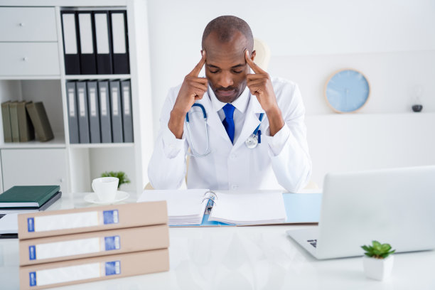 家庭医生的照片黑皮肤专业的家伙写处方病人基础期刊手指在太阳穴工作日夜累穿白色的实验室大褂坐椅子现代办公室诊所室内图片下载