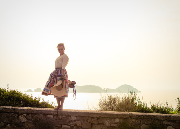 快乐的年轻女子戴着帽子，穿着优雅多彩的裙子，赤脚站在海边的一面墙在夕阳意大利庞扎岛。武器扩散。图片下载