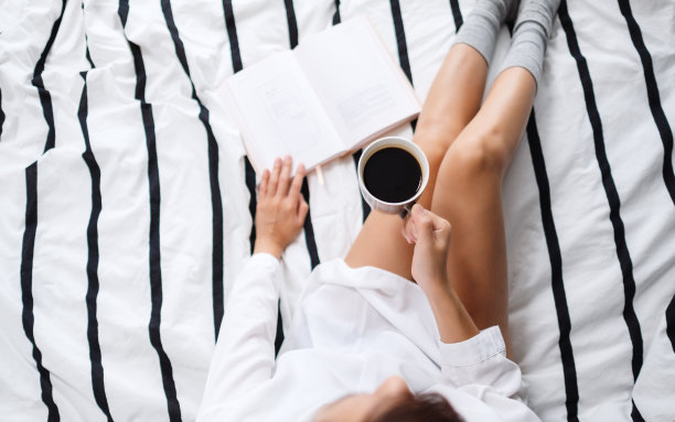 早上，一个女人坐在舒适的白色床上看书，喝着热咖啡图片下载