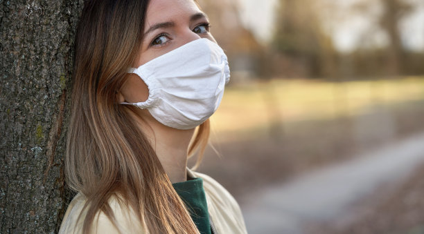 年轻女子戴着白色棉病毒口罩在公园里，靠在这里的树上图片下载