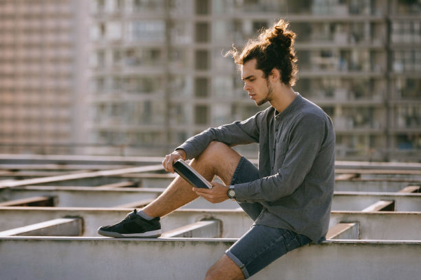 男人坐在屋顶上看书和喝咖啡茶，喝纸杯。背景的城市风光。城市的生活方式图片下载