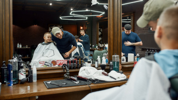 朋友。年轻的理发师和可爱的小男孩在理发店看着镜子微笑。儿童理发沙龙图片下载