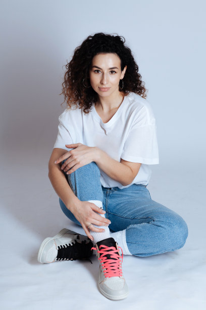 迷人的卷曲的黑发，白色t恤，牛仔裤和运动鞋在一个灯光工作室的背景图片下载