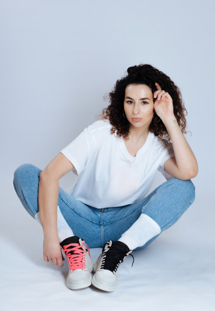 迷人的卷曲的黑发，白色t恤，牛仔裤和运动鞋在一个灯光工作室的背景图片下载