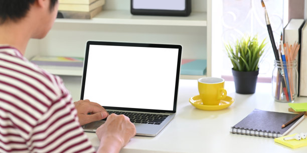 聪明的人在白色空白屏幕的笔记本电脑上打字的背影，放在白色的办公桌上，周围是咖啡杯，盆栽植物和文具。图片下载