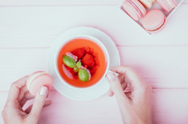 女性的手拿着一杯白色的覆盆子茶和粉红色的杏仁饼干在一个淡粉色的背景。俯视图图片下载