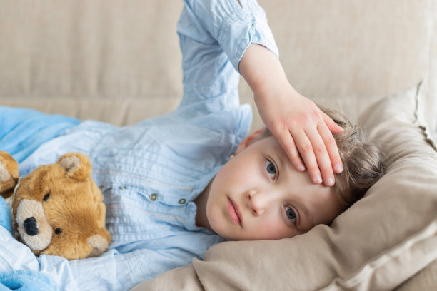 女孩躺在家里的米色沙发上，手里拿着一只泰迪熊，用手测量额头上的温度。生病的孩子发烧流感图片下载