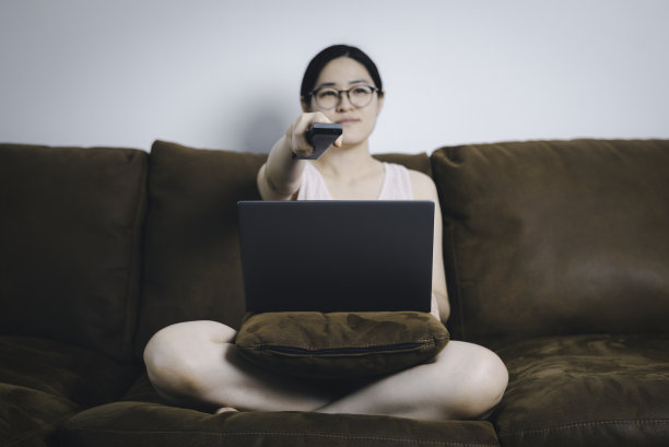 亚洲女性在沙发上用笔记本电脑工作，拿着遥控器搜索电视节目。在家进行概念工作图片下载