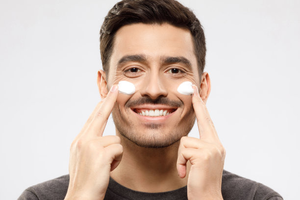 英俊的年轻人看着镜头，用手指触摸脸颊，涂面霜，感觉满意的美容常规，孤立在灰色的背景。皮肤护理人图片下载