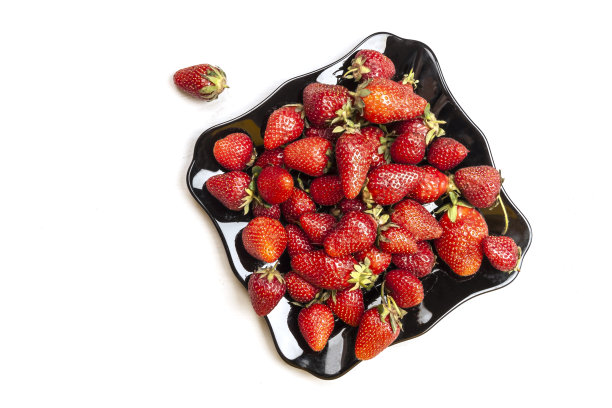 俯视图新鲜美味的草莓在黑色光泽的盘子和几个草莓分别在白色的背景。夏天的时间概念图片下载