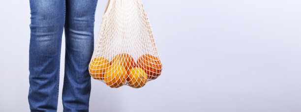 穿着蓝色牛仔裤的年轻女子手持有各种柑橘灰色背景的网袋。概念零浪费，环保。本空间图片下载