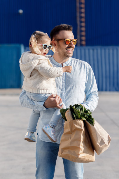 一个快乐的父亲肩上扛着一个小女儿走在停车场里，手里拿着装着新鲜蔬菜和购物的纸袋。图片下载