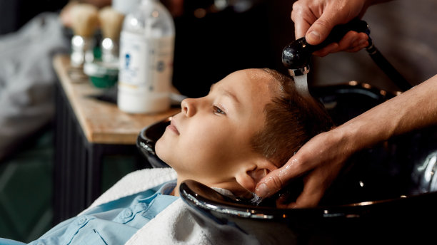 对孩子理发。一个男性理发师在理发店里洗可爱的小男孩的头发的特写照片。孩子们在理发店图片下载