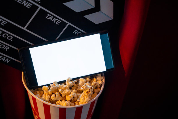 手机与空白色明亮的屏幕爆米花桶和电影拍板。在互联网手机上播放电视的概念。图片下载