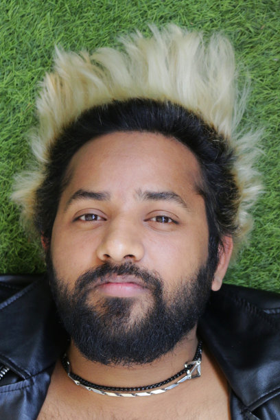 一个年轻英俊的印度男子穿着皮夹克，20出头看着镜头微笑着，他留着短胡子，染过的头发凌乱地散布在周围。一个来自印度德里的英俊男子的特写，他戴着项链躺在外面的草地上图片下载