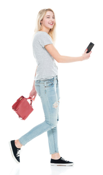 z一代的年轻女性穿着裤子，拿着公文包，用着智能手机走在白色背景前图片下载