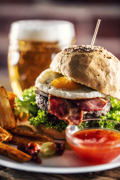 汉堡配芝麻面包、煎蛋和培根、沙拉、土豆块和辣椒酱，背景是生啤酒。图片下载