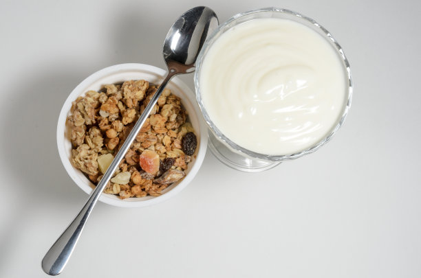 酸奶放在一个漂亮的玻璃碗里，旁边放一盒格兰诺拉麦片和一把勺子。图片下载