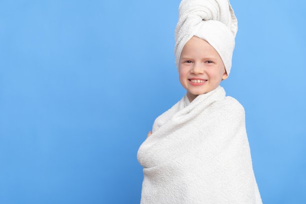 可爱的小白人女孩的肖像包裹在白色的毛巾与浴巾在头上的蓝色工作室背景。水疗程序，沐浴纺织品图片下载