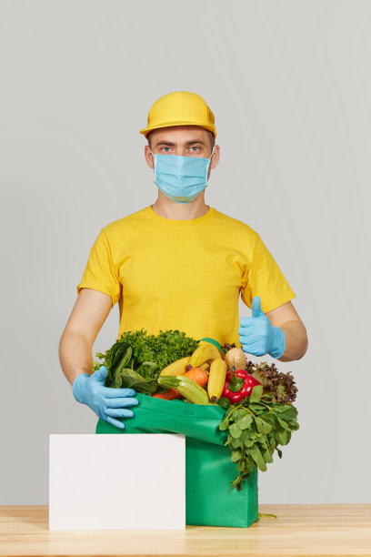 网上购物的概念。男快递员身穿黄色制服，戴着防护口罩和手套，带着一个装着新鲜水果和蔬菜的食品盒。冠状病毒隔离期间送餐到家图片下载