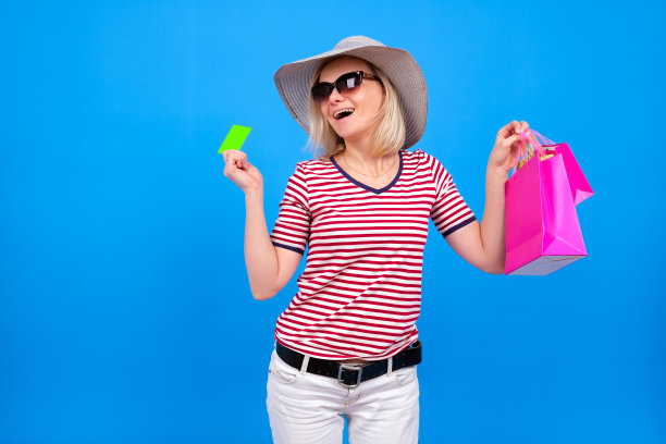 兴奋的金发白人女孩在夏天的帽子拿着紫色的购物袋和显示绿色信用模拟卡，孤立在蓝色的背景。Copyspace。夏季购物和销售概念图片下载