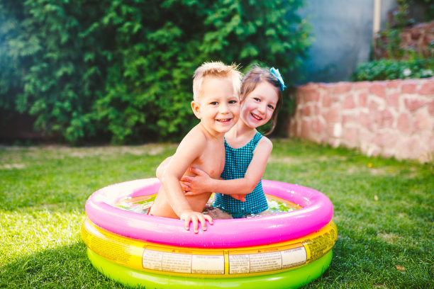主题是儿童的暑假。两个白种小孩，弟弟和妹妹，坐在院子里的绿油油的草地上，穿着游泳衣坐在一个圆圆的水池里，快乐幸福的微笑着图片下载