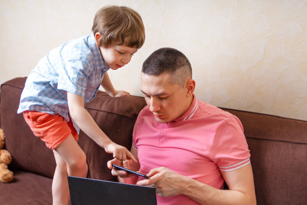 快乐的年轻父亲坐在电脑上使用笔记本电脑放松与学龄前的儿子一起玩，微笑的父亲和小男孩孩子享受周末在家休息在沙发上忙着小玩意。在线教育图片下载