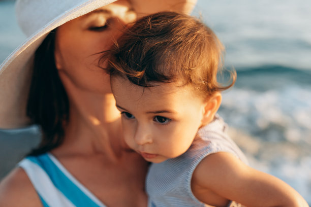 近距离拍摄的照片，母亲亲吻她的女儿在海上的背景。妈妈亲吻她的孩子。白种女性戴着帽子和孩子在海洋摆姿势图片下载