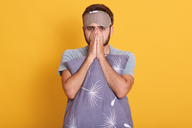 室内照片，一名留着胡须的男子，穿着t恤，蒙着眼睛，抱着灰色枕头，双手合拢，捂着脸，靠在黄色的墙壁上。图片下载