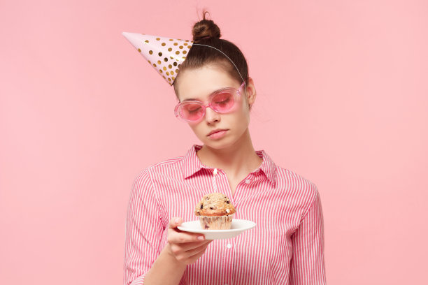 年轻的生日女孩戴着眼镜和帽子，拿着装有节日纸杯蛋糕的盘子，带着悲伤的脸看着它，厌倦了派对，孤立在粉红色的背景上图片下载