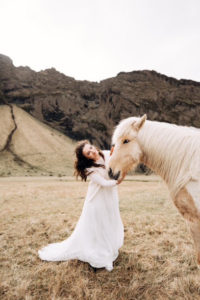 在一片黄色的草地上，新娘抚摸着一匹有着奢华鬃毛的奶油色马的脸，旁边是一座岩石山。目的地冰岛婚礼照片会议与冰岛马。图片下载