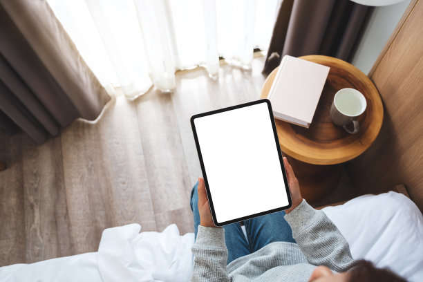 一个女人坐在舒适的白色床上，手里拿着黑色平板电脑和空白的桌面白色屏幕图片下载
