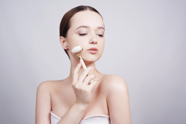 肖像照片的一个年轻女子看起来轻松使用而使用天然白色石英面部滚轮。美丽的脸。妇女做脸部按摩与玉面部滚轮为水疗皮肤护理治疗在家。图片下载