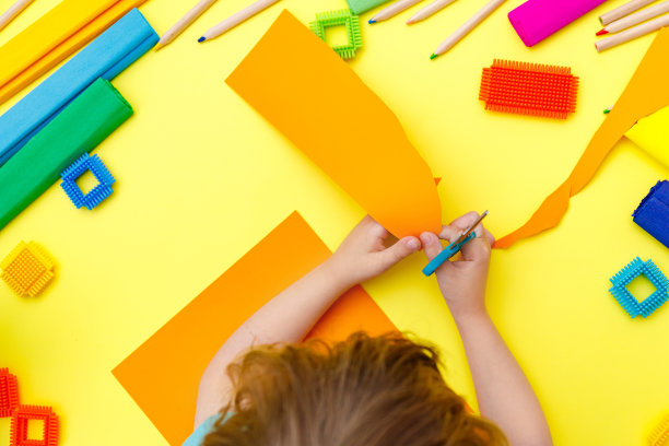 孩子在桌子上用剪刀剪橙色的纸做工艺品图片下载