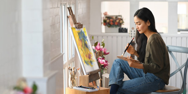 美丽的女人用画笔在画布上绘画，而坐在椅子上现代和舒适的艺术工作室的窗户作为背景。工作女性艺术家的概念。图片下载