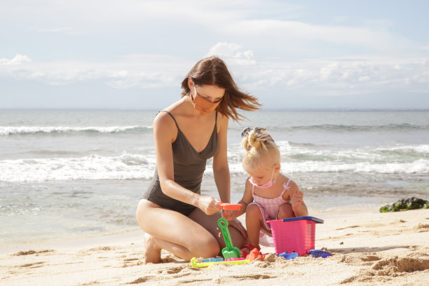 新冠肺炎疫情危机后的暑假。可爱的小女孩和她的妈妈戴着防护面具在沙滩上玩沙滩玩具。图片下载