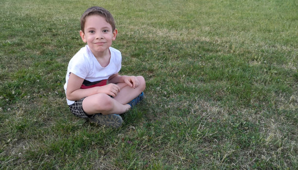 一个5岁的小男孩，对着镜头微笑，盘腿坐在草地上，草地上有绿色的，有点干的草。孩子穿着白色t恤，黑白格子短裤和凉鞋。水平横幅，复制空间图片下载