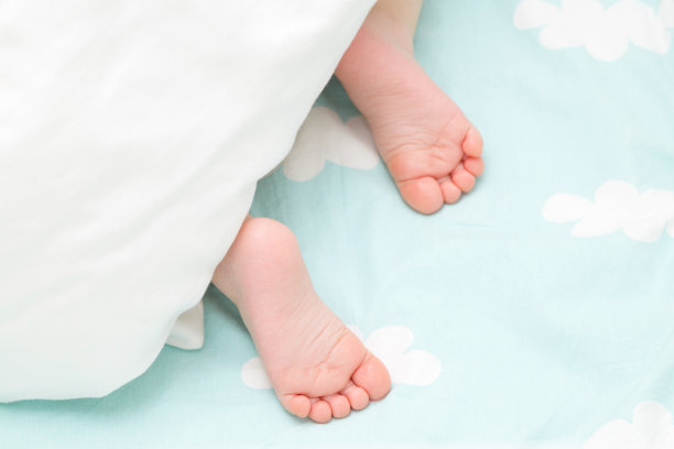 婴儿赤脚躺在床上。脚伸出白色毯子。宝宝睡觉。特写镜头。蓝色的床单和云彩。图片下载