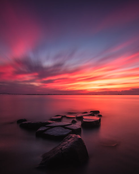 美丽的日落照亮了天空，平静的水包围着岸边的岩石。纽约火岛图片下载