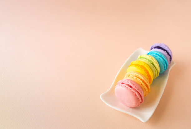 6个不同颜色的手工蛋白杏仁饼干放在白色的长方形盘子上，照片的右边是极简的构图。饼干的颜色有粉色、蓝色、浅绿色、黄色、紫色和橙色。物体在浅粉色的背景与纸图片下载