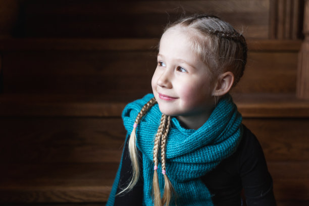 漂亮的白人小女孩坐在木楼梯上，金色的头发编成辫子，黑色的眼睛戴着蓝色的围巾，看着窗户微笑着图片下载