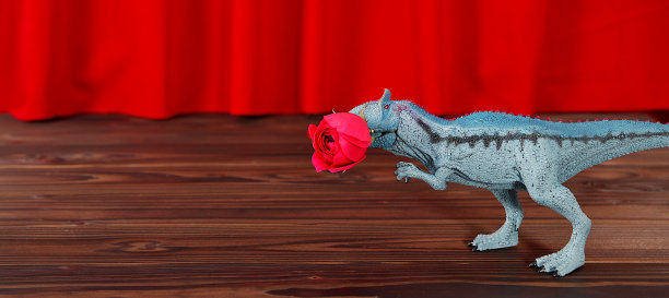 舞台上的塑料恐龙。红色剧场幕布背景上插着红玫瑰的龙玩具。有趣的情人节贺卡。创造性的最小概念。复制空间，文本的自由空间。图片下载