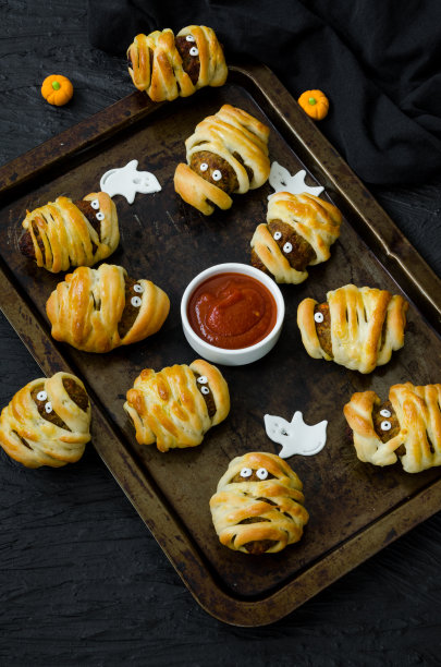 万圣节木乃伊-肉丸包裹在生面团和辣番茄酱在一个旧烤盘上在黑色的背景。图片下载
