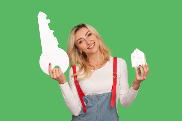 快乐的金发女人的肖像在时尚的工装裤举起大钥匙和纸房子，看着相机与牙齿微笑图片下载