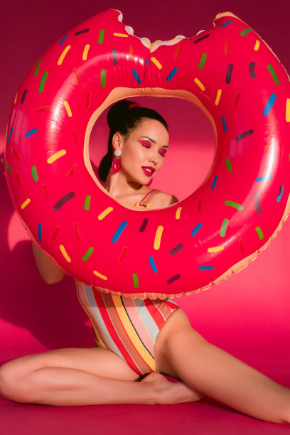 肖像苗条丰满的身体性感诱人的性感美丽的女人在复古泳衣微笑，闭着眼睛坐在看通过油炸圈饼形状的浮动孤立在粉红色的背景洞图片下载
