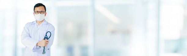 在医院的横幅背景下，亚洲男医生戴着防止细菌的口罩，站在模糊的内部走廊前。医院有效工作理念。图片下载