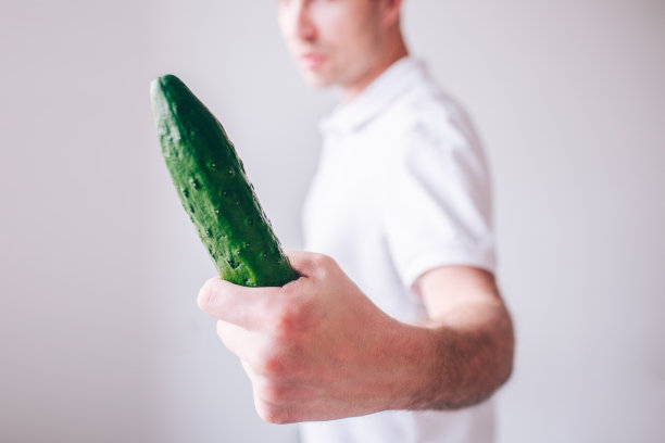 穿白衬衫的年轻人在背景中显得很孤立。那家伙看着长长的黄瓜手里。剪切视图模糊散焦背景。图片下载