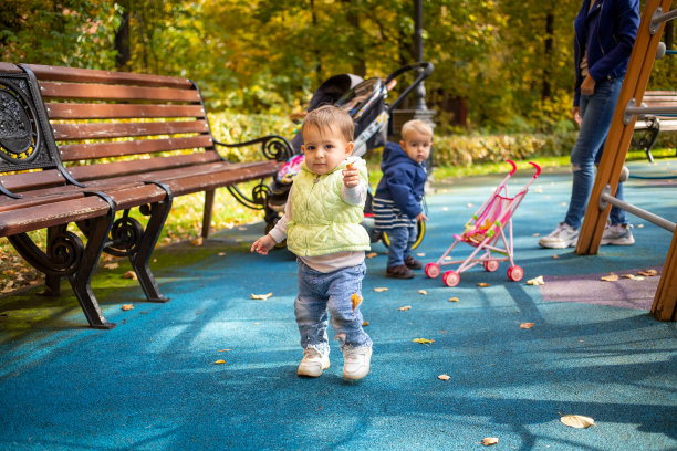 一个可爱的小婴儿在公园里玩耍，看着镜头，伸出一片黄色的树叶。另一个婴儿站在后面看着镜头，有一个婴儿车和妈妈。软焦点，背景模糊。图片下载