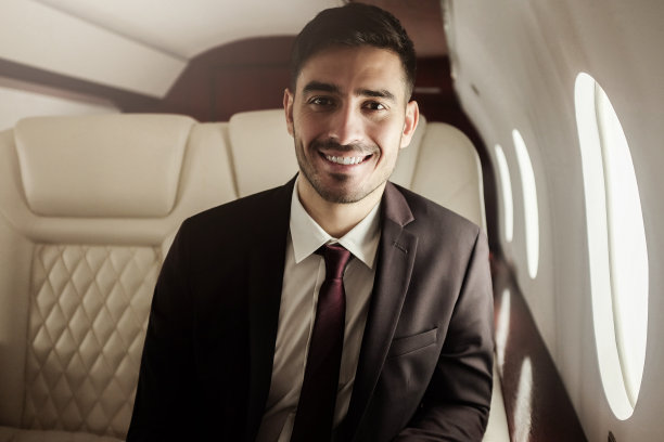 这是一个年轻帅气的商人在私人飞机上的照片，他穿着西装，打着领带，坐在靠窗的座位上，心情愉快，充满自信图片下载