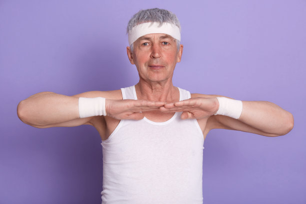 照片中，一位精力充沛、面容俊美的退休老人伸展着身体，孤立地站在工作室里的淡紫色背景下，举着手臂直视镜头，穿着白色衬衫，头戴发带。健康的概念。图片下载